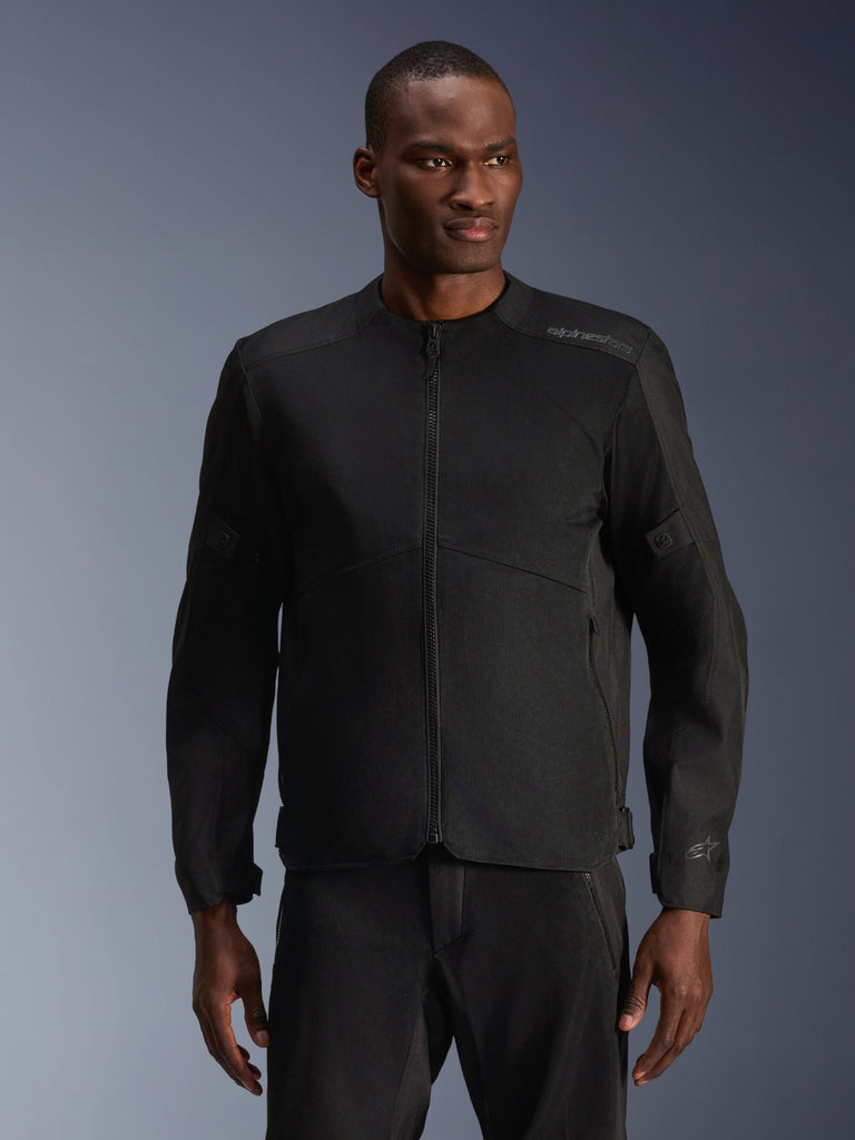 Modelo masculino a usar Casaco Alpinestars C-1 Air em Preto, casaco de motociclismo contemporâneo com amplos painéis de malha técnica e fecho de correr frontal integral, combinado com calças de condução pretas a condizer, estética urbana e de deslocações diárias