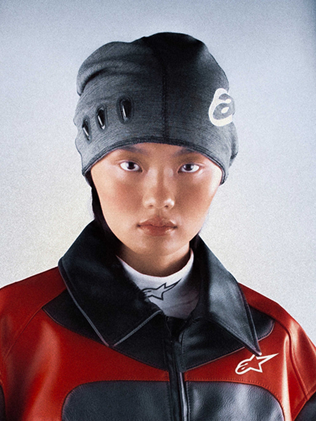 Alpinestars RSRV Onyx Beanie, gorro unissexo cinzento-escuro com recortes ovais e design branco, usado com casaco vermelho e preto de colarinho alto