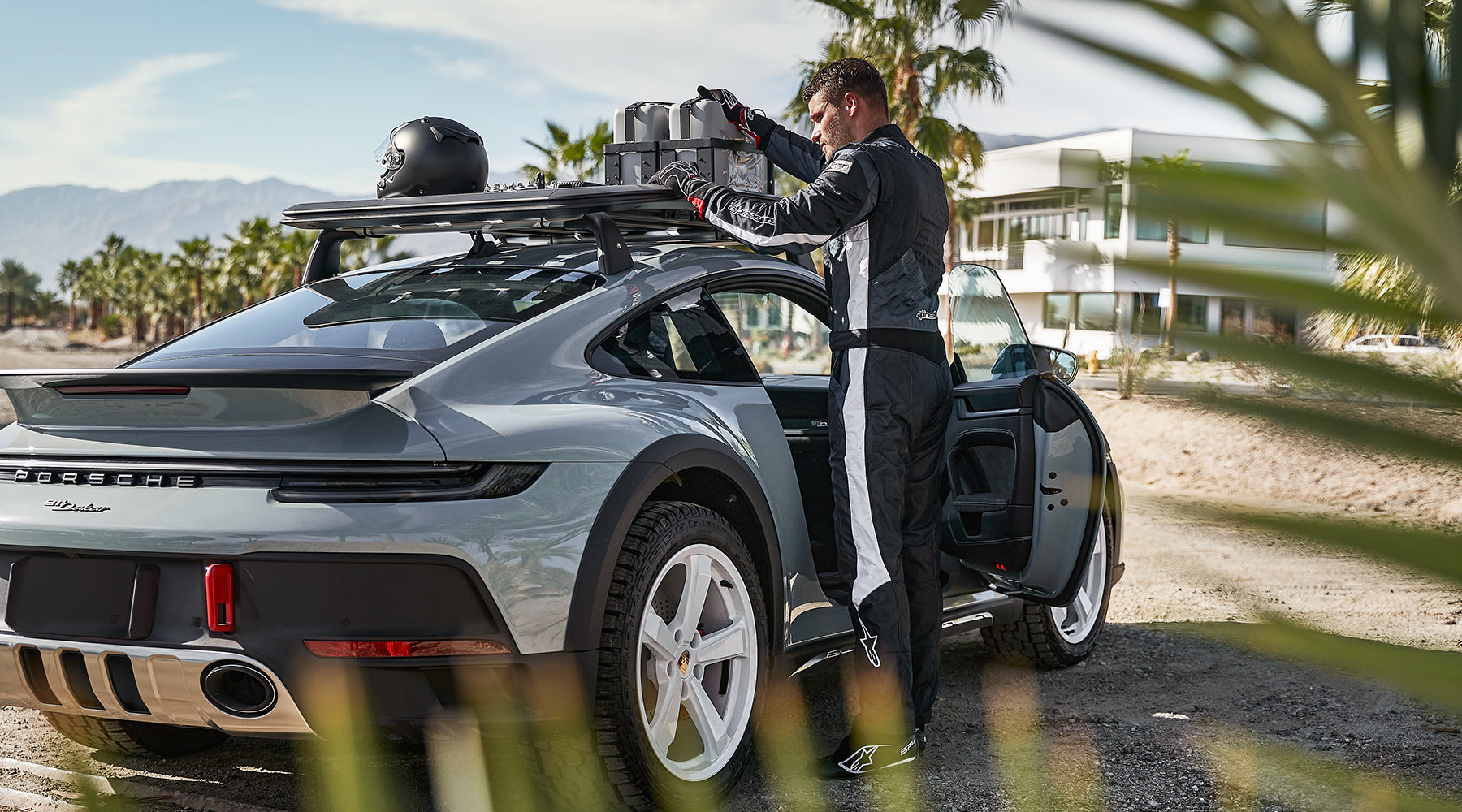 Alpinestars GP Pro Comp V2 FIA Suit, asphalt and white auto racing suit, worn by a driver loading a Porsche 911 Dakar.