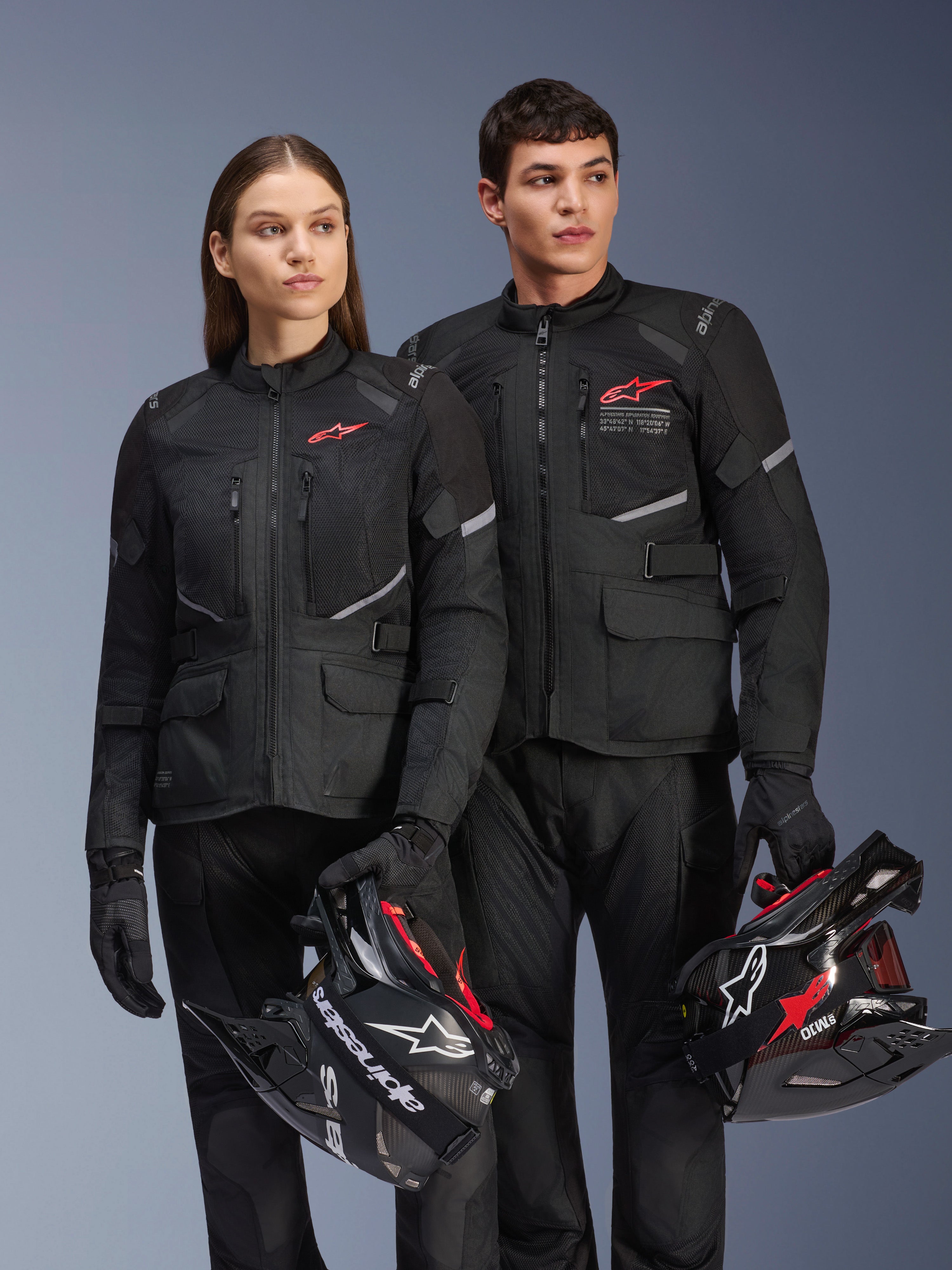 Homem e mulher a usar casacos Alpinestars Andes Air Drystar®, equipamento de touring de aventura preto com painéis de malha e detalhes refletores, calças e luvas pretas a condizer, a segurar capacetes de motocross, fotografia de estúdio de vestuário profissional de motociclismo